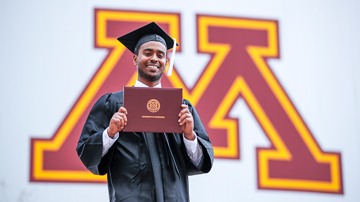 Student holding diploma smiling at camera, block M logo in background.